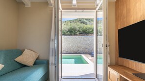 Une porte coulissante ouverte donnant sur une piscine, un canapé turquoise agrémenté de coussins et un meuble de rangement en bois abritant un téléviseur.