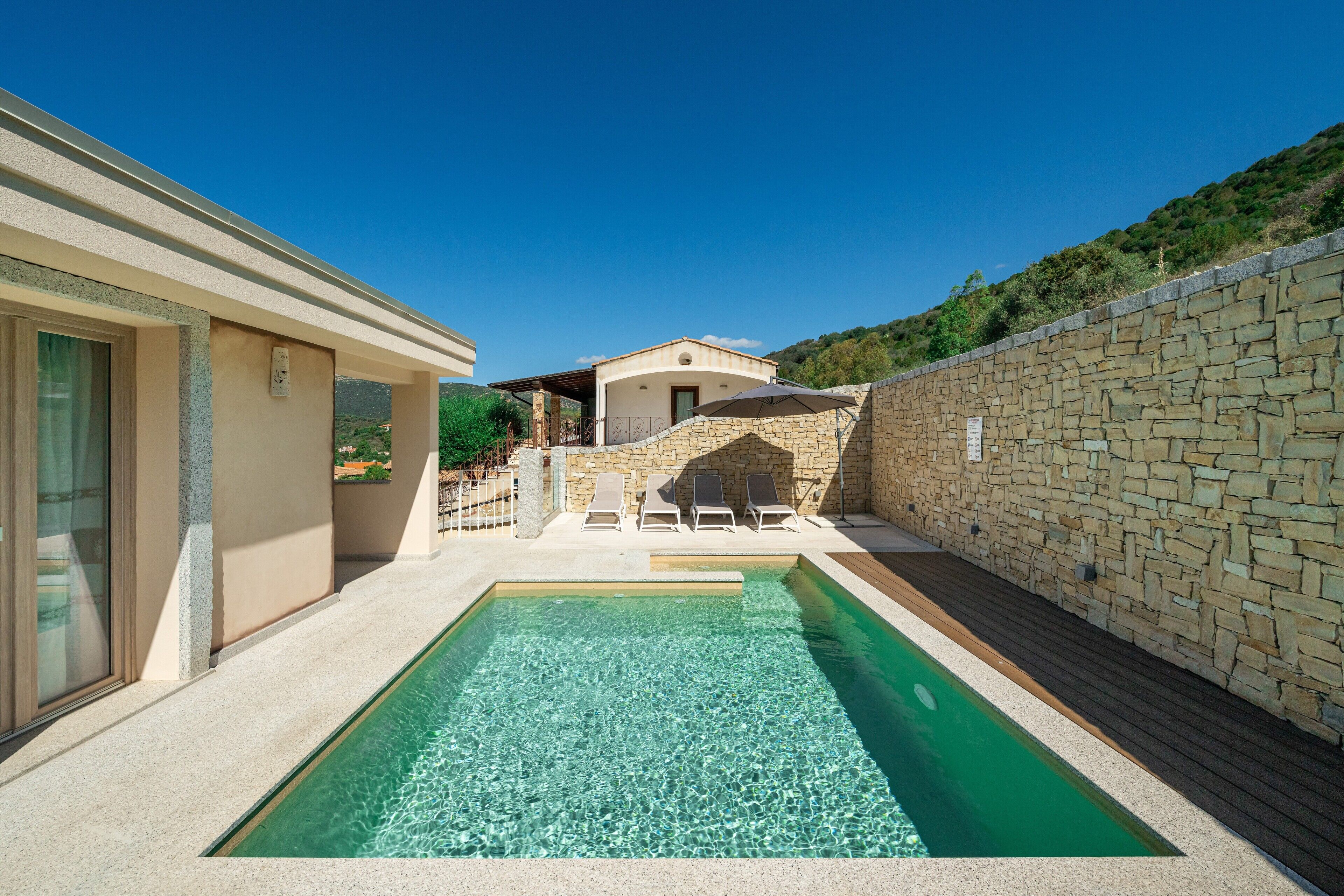 Une piscine avec des chaises longues, un mur en pierre et un bâtiment doté d’un patio couvert.