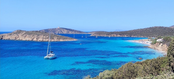 A coastal landscape with clear blue waters, a sailboat, and rocky hills.
