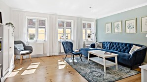 A living room with a blue sofa, a grey armchair, a white coffee table, and a patterned rug. There are two windows with curtains, a radiator, and framed pictures on the wall. - 3 bedroom lovely home in Faaborg (Faaborg)