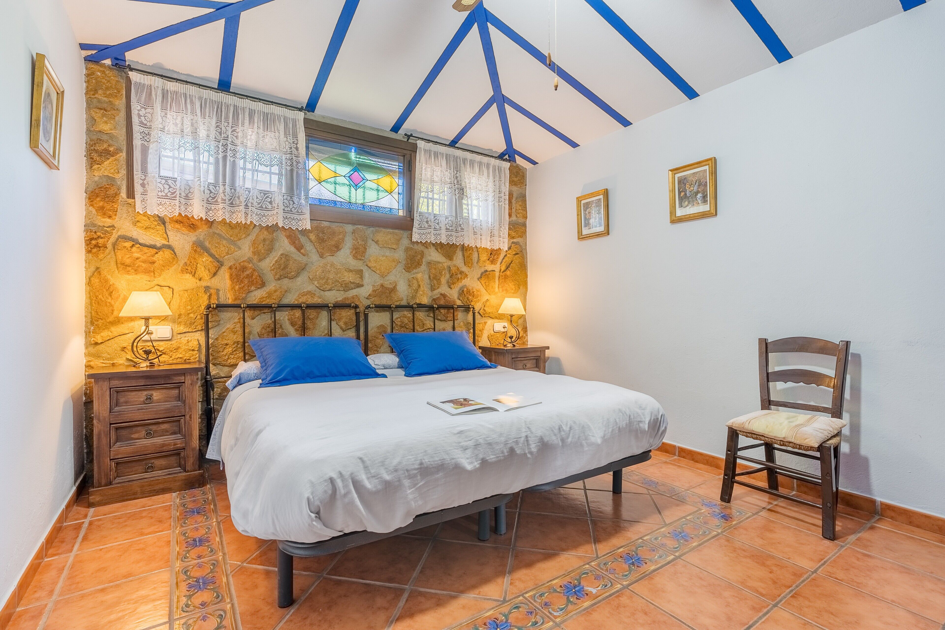 Une chambre avec un mur en pierre, un lit avec des oreillers bleus, une chaise et une table de chevet avec une lampe.