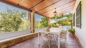 Un patio techado con una mesa y sillas, paredes de piedra y vistas a zonas verdes y una piscina.
