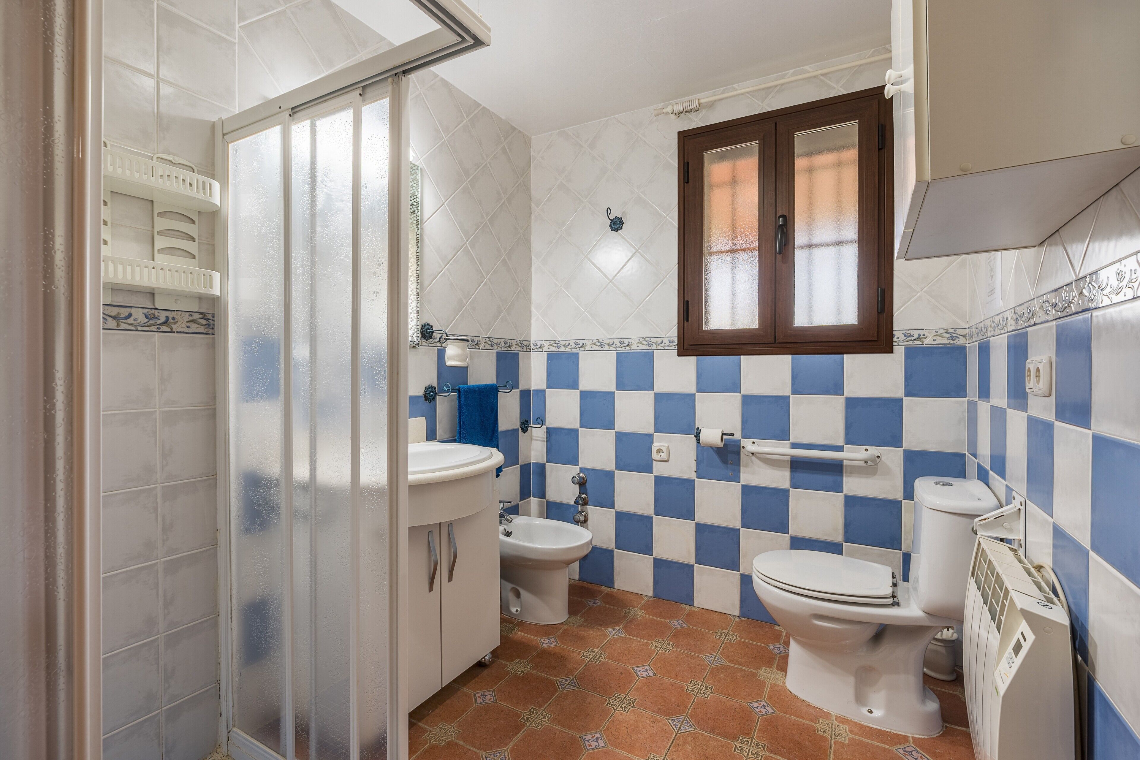 A bathroom with a toilet, bidet, and sink, tiled in blue and white, with a window above the toilet.