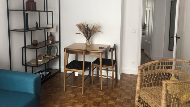 A room with a wooden floor, a blue sofa, a dining table with chairs, a wicker chair, a black-framed bookshelf, and a small wall-mounted picture frame. - COSY Belleville briapartment (Paris)
