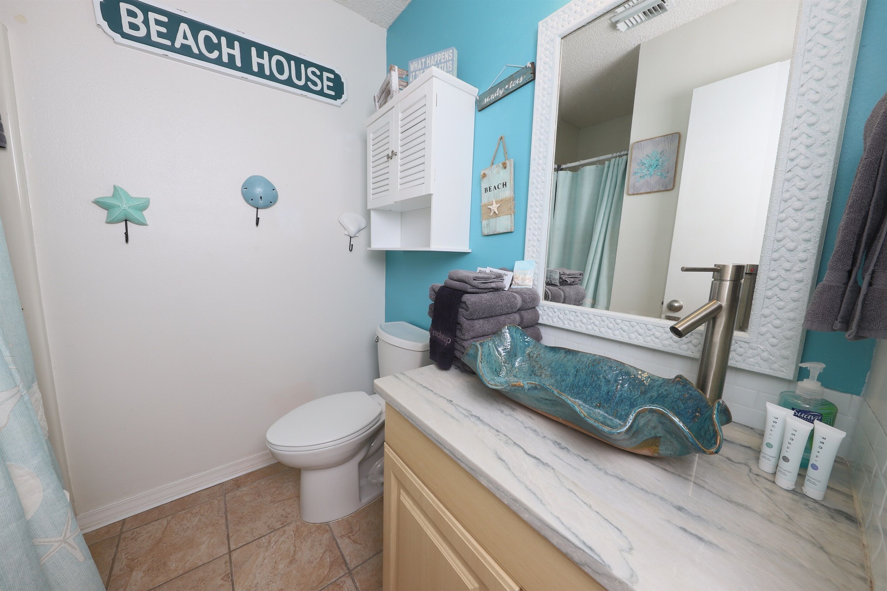 Een badkamer met toilet, een wastafel met een blauwe keramieken kom en een spiegel boven de wastafel. Er is een handdoekhouder met handdoeken en een bordje met de tekst 'BEACH HOUSE'.