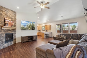 A spacious living area with a fireplace, a ceiling fan, a kitchen in the background, and large windows. - Woodland Haven | On Golf Resort | 10 min Bearizona (Williams)