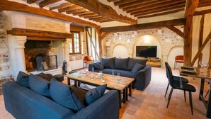 A rustic living room with stone walls, wooden beams, a fireplace, a television, and a dining area with a table and chairs.