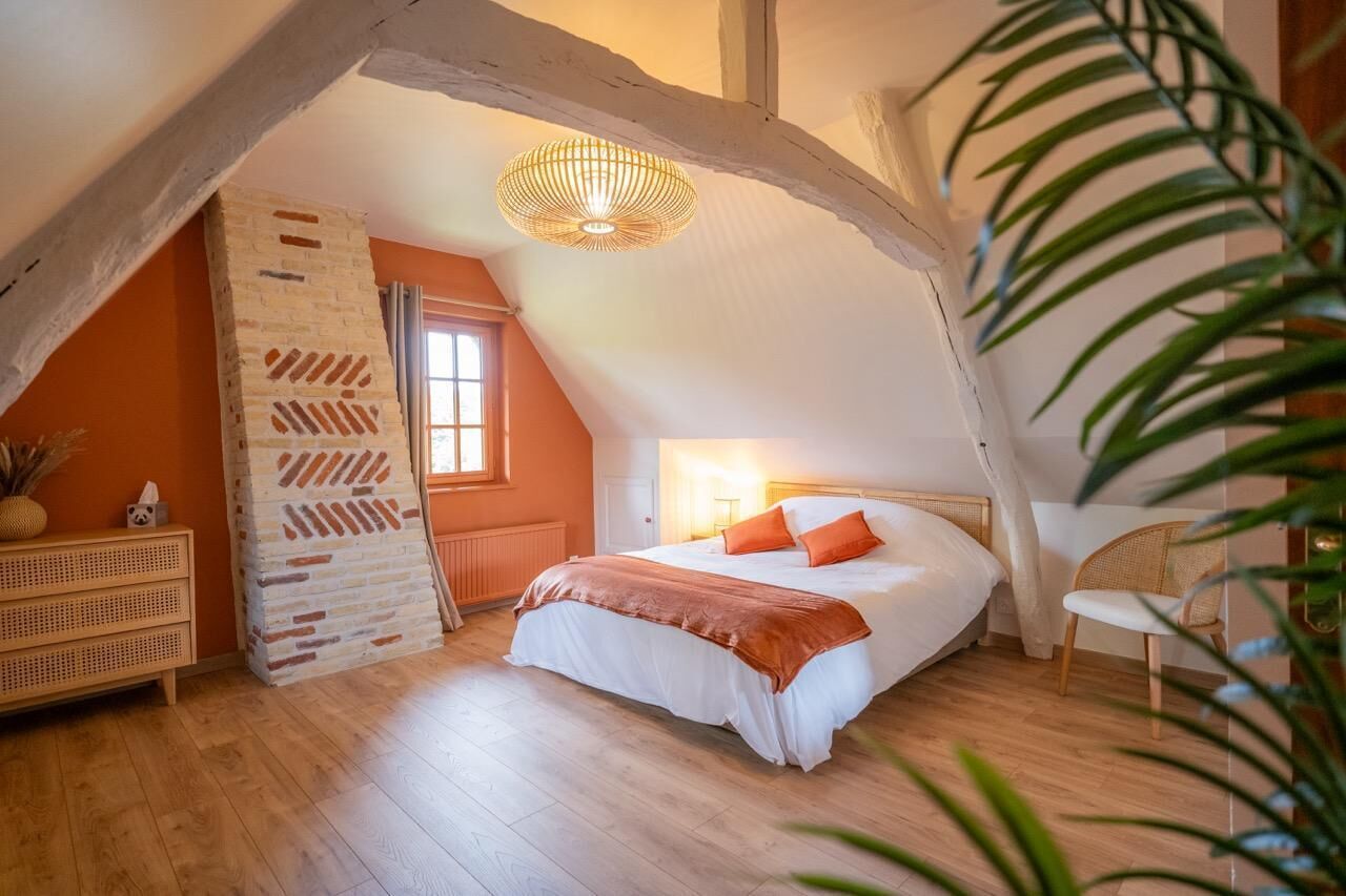 A bedroom with a sloped ceiling, a bed with white bedding and orange pillows, a wicker chair, and a hanging light fixture.