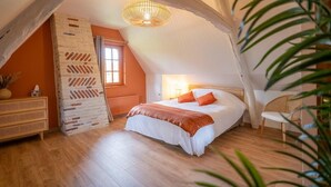 A bedroom with a sloped ceiling, a bed with white and orange bedding, a wicker chair, and a hanging light fixture.