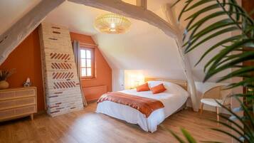 A bedroom with a sloped ceiling, a bed with white bedding and orange pillows, a wicker chair, and a hanging light fixture.