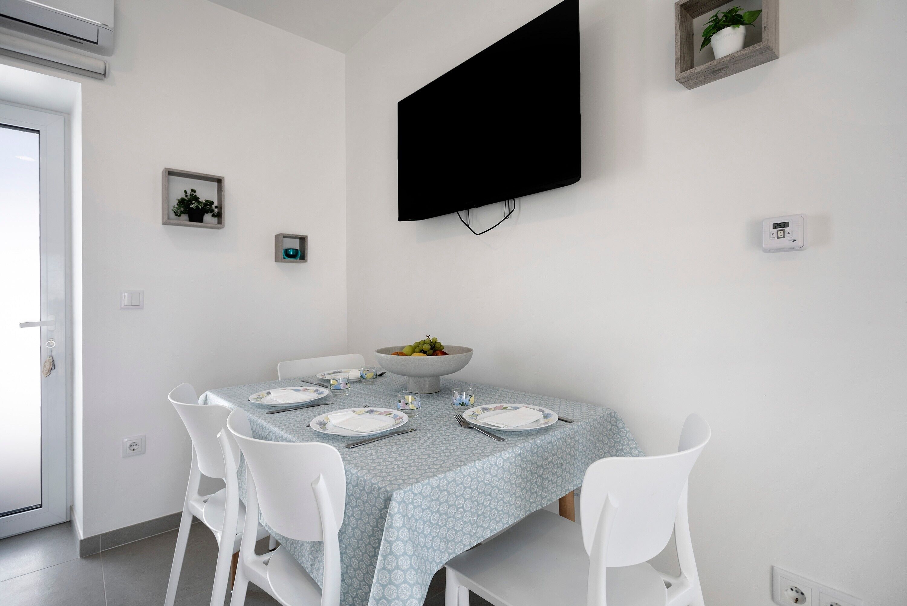 Een eetruimte met een tafel voor vier personen, witte stoelen, een wandgemonteerde flatscreen televisie en een kleine potplant.