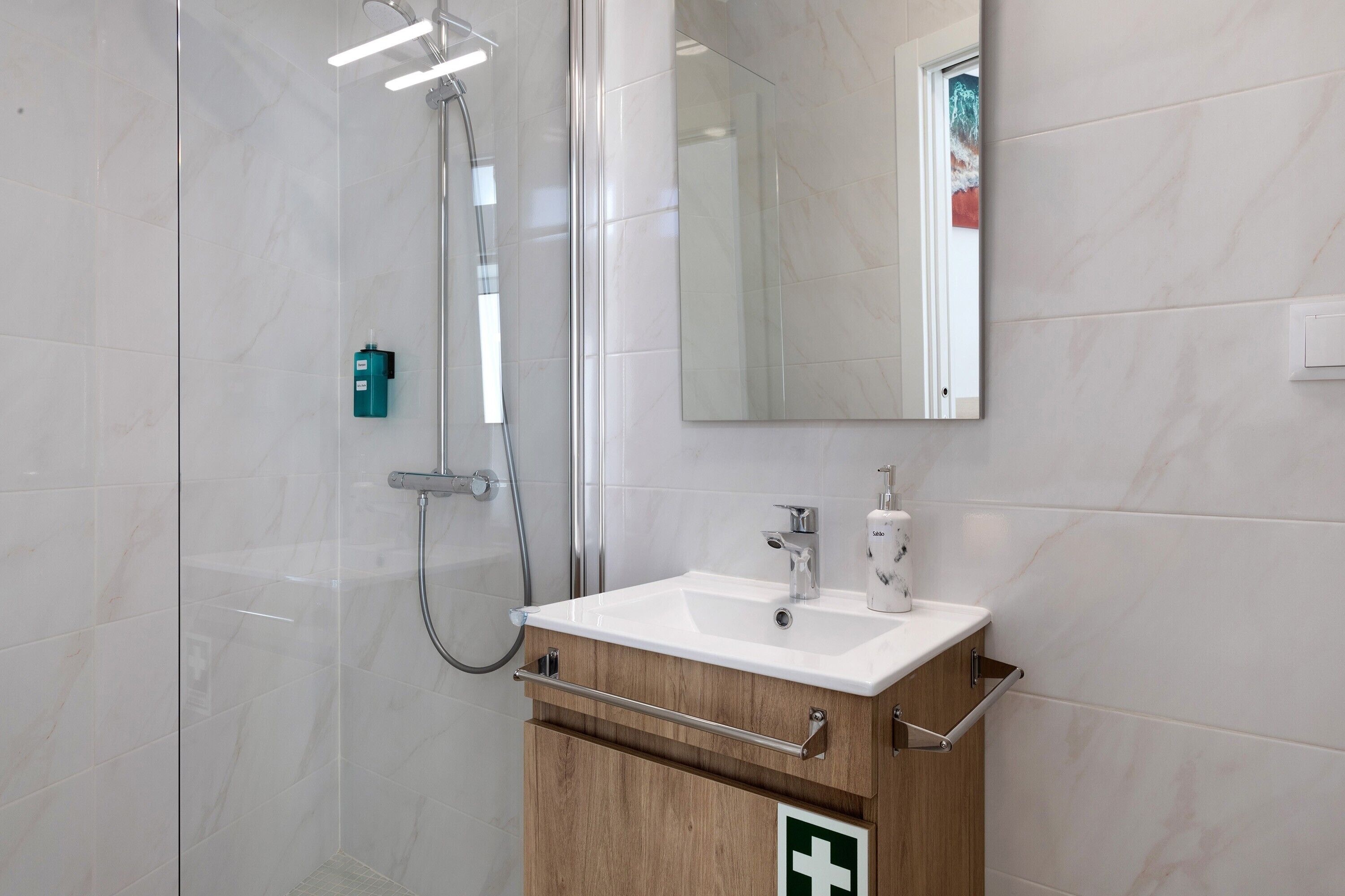 Une salle de bain moderne avec une douche à l’italienne en verre, un lavabo blanc avec un miroir au-dessus, et un meuble en bois sur lequel est apposé un panneau vert indiquant les consignes de sécurité.