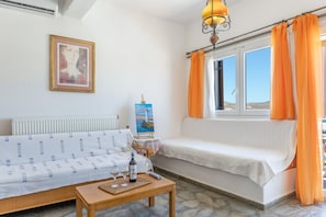 A room with a sofa, a table with a bottle and glasses, a painting on the wall, and a window with curtains.