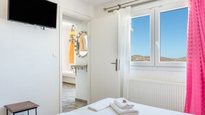 A hotel room with a bed, a television mounted on the wall, a window with curtains, and a bathroom visible through an open door. - Apartment 'Agnadi' with Sea View, Balcony and Air Conditioning (Serifos)