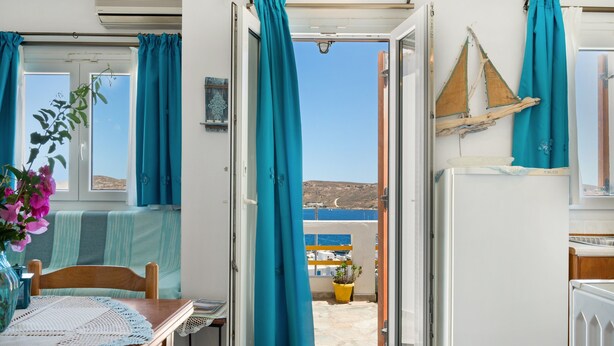 Eine geöffnete Tür, die auf einen Balkon mit Meerblick führt, mit einem Holzstuhl, einem Tisch mit weißer Spitzenleinentischdecke und einem Blumentopf.