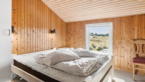A bedroom with a wooden sloped ceiling, a bed with white bedding, a nightstand, a wall-mounted lamp, and a view of the outdoors through a glass door. - Pet friendly home in Thisted with sauna (Thisted)