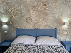Uma cama com cabeceira azul, almofadas brancas e um edredom com padrão de árvores. Há duas luminárias fixadas na parede, de cada lado da cama.