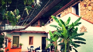 A courtyard with a stone building, outdoor seating, and a large banana plant. - Triple room with shared pool and kitchen, Wi-Fi and air conditioning (Gonnesa)
