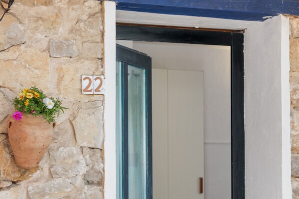 A stone wall with a number plaque, a hanging flower pot, and a doorway leading to a room.