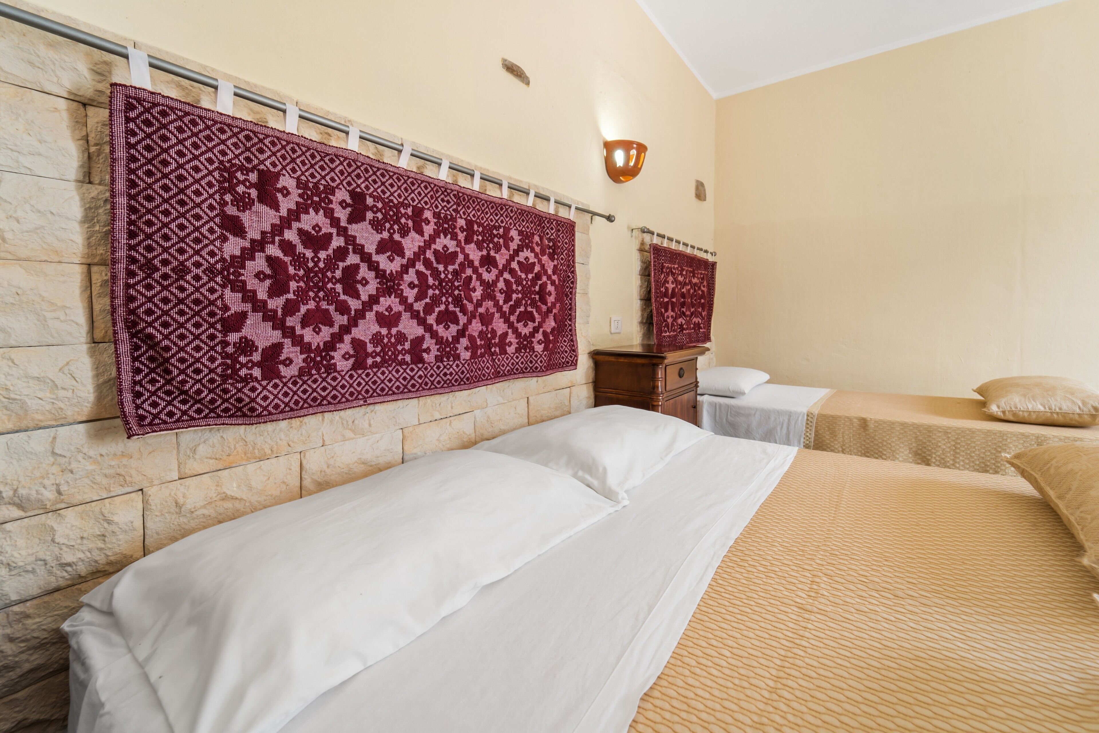 A hotel room with a stone wall, a large patterned rug, two beds with white and beige bedding, and a small wooden nightstand.