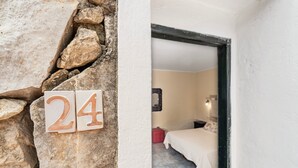 A stone wall with a number '24' sign, a bedroom with a bed and a nightstand, and a white wall. - Double room with shared pool and kitchen, Wi-Fi and air conditioning (Gonnesa)