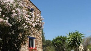A stone building with a window and a small balcony, surrounded by lush greenery and flowering plants. - Double room with shared pool and kitchen, Wi-Fi and air conditioning (Gonnesa)