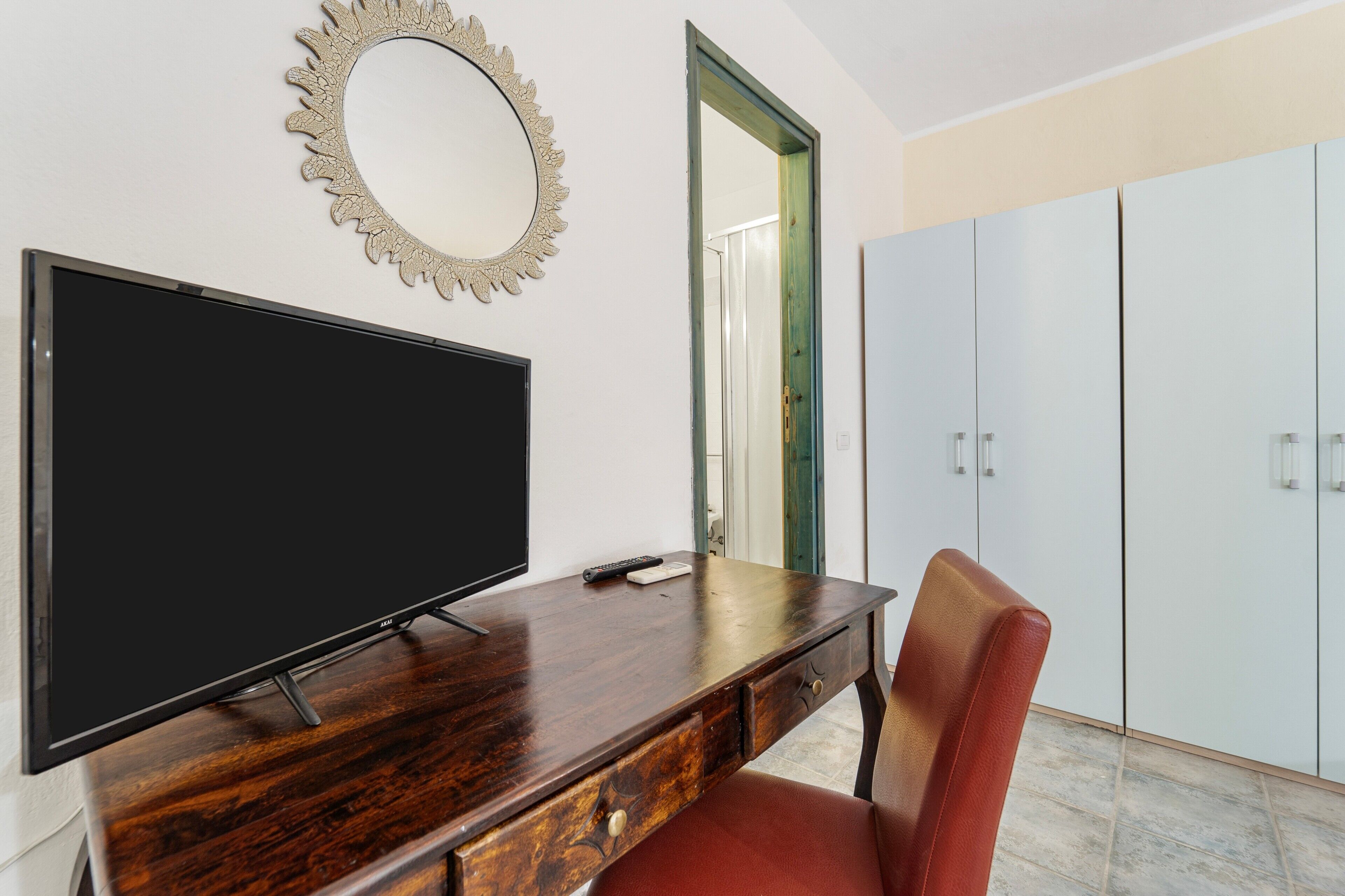 Une pièce comprenant un téléviseur à écran plat posé sur un bureau en bois, une chaise rouge, un miroir décoratif et une armoire.