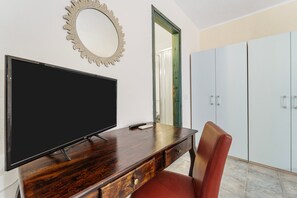 Quarto com televisão de tela plana sobre uma escrivaninha de madeira, uma cadeira vermelha, um espelho decorativo e um guarda-roupa.