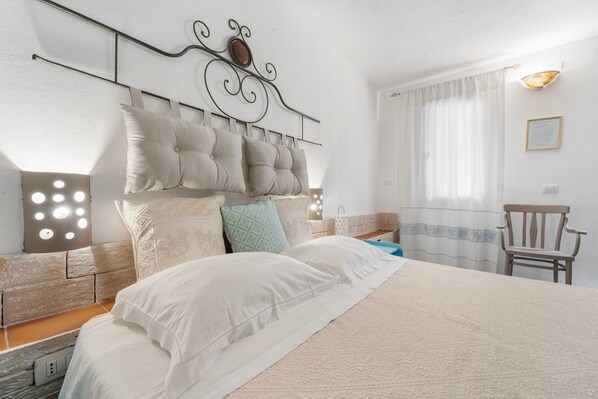 A neatly made bed with a headboard, a bedside table with a lamp, and a chair in a room with white walls and a window with curtains. - Double room with shared pool and kitchen, Wi-Fi and air conditioning (Gonnesa)