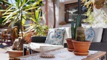 Um pátio com uma mesa, dois cactos em vasos, um jogo americano de fibra natural e um sofá de vime com almofada azul.