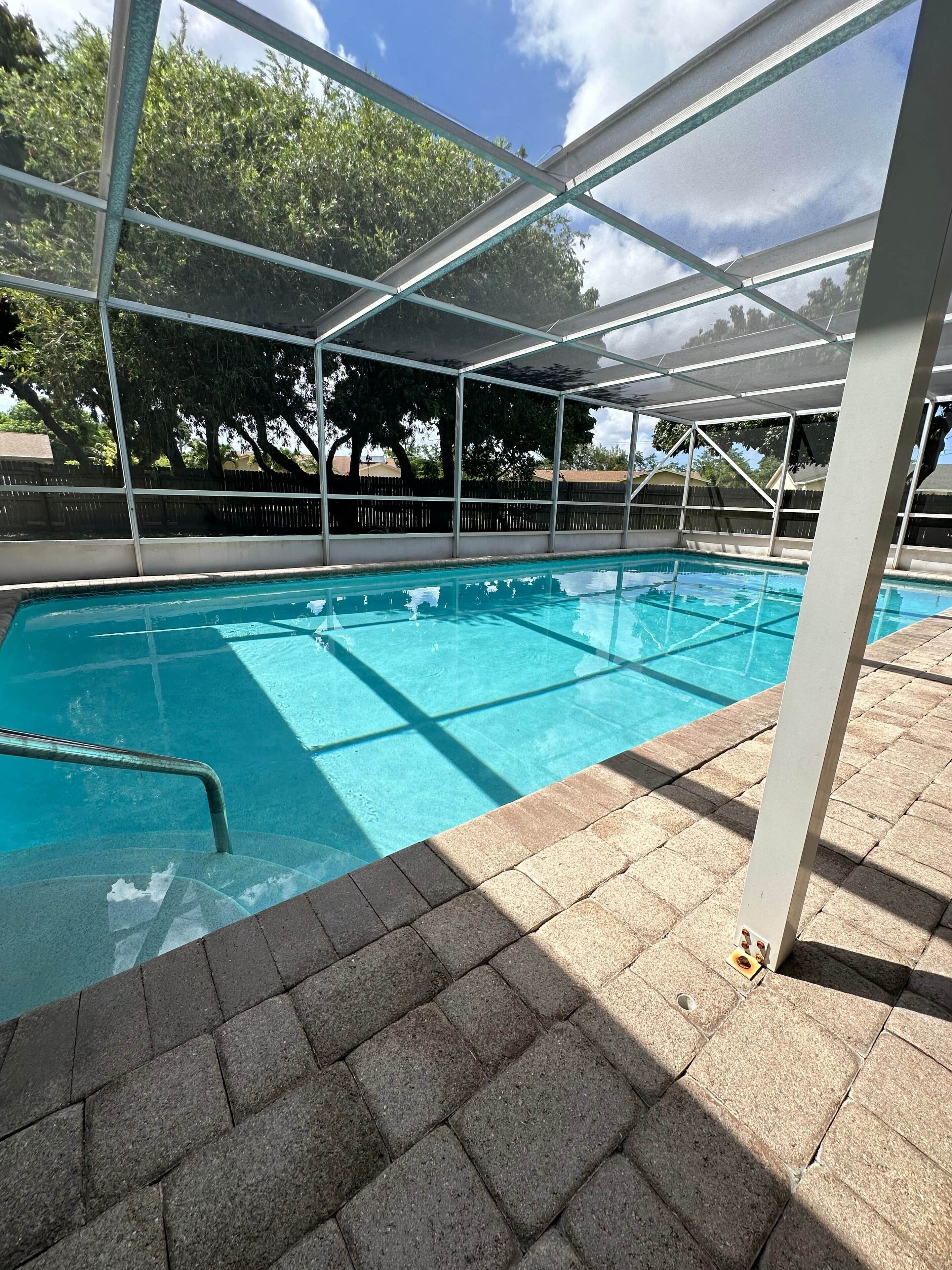 A clear swimming pool with a covered area, surrounded by a brick deck and trees in the background.