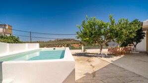 Una piscina con un deck blanco, un árbol y una mesa con sillas en un entorno exterior soleado.