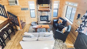 A cozy cabin interior with a stone fireplace, a flat-screen TV mounted above it, a sofa, a coffee table, and a dining area with chairs and a wooden table.