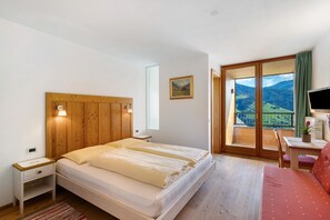 A bedroom with a wooden headboard, a bed with white bedding, a nightstand, a desk with a chair, a TV, and a balcony with a view of the countryside.