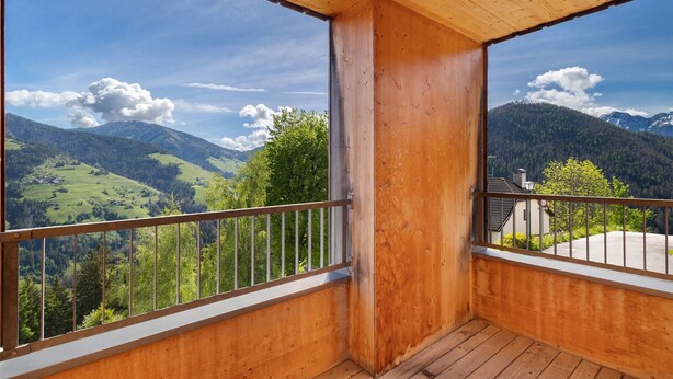 Un balcón de madera con barandilla metálica, que ofrece vistas a colinas y montañas verdes.