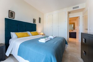 Een net opgemaakt bed met een blauw-witte spreidek, een donkerblauw hoofdbord en twee kussens. Twee ingelijste foto's hangen aan de muur, een nachtkastje met een lamp en een garderobe met een spiegel.