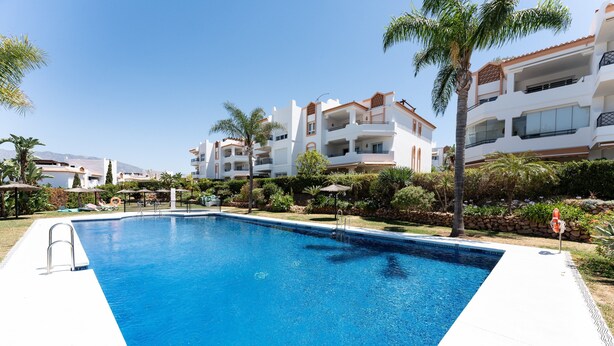 Una piscina con agua cristalina azul, rodeada por un solárium blanco, palmeras y un edificio con balcones al fondo.