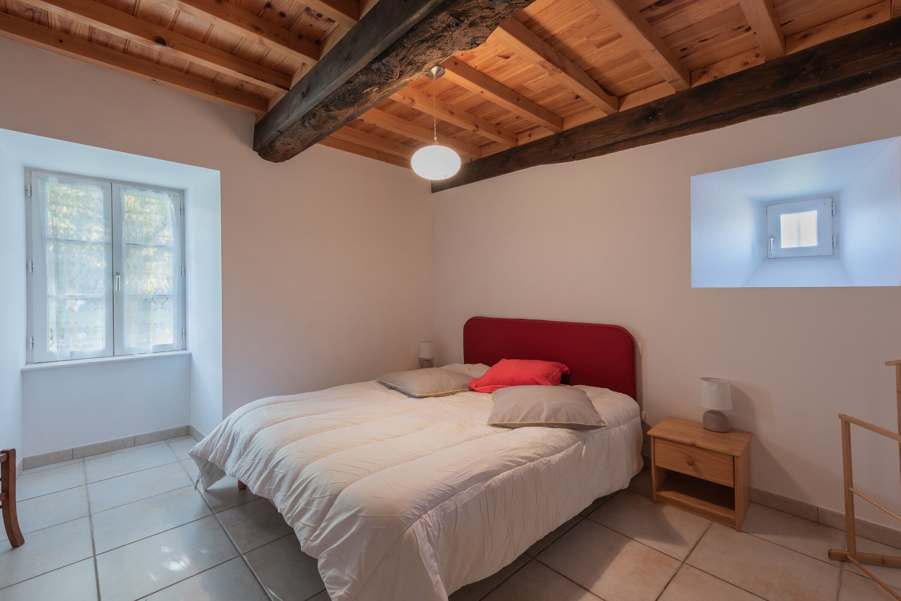 Een slaapkamer met een houten plafond, een bed met witte beddengoed en een rode hoofdbord, een nachtkastje met een lamp, en een raam met uitzicht op bomen.