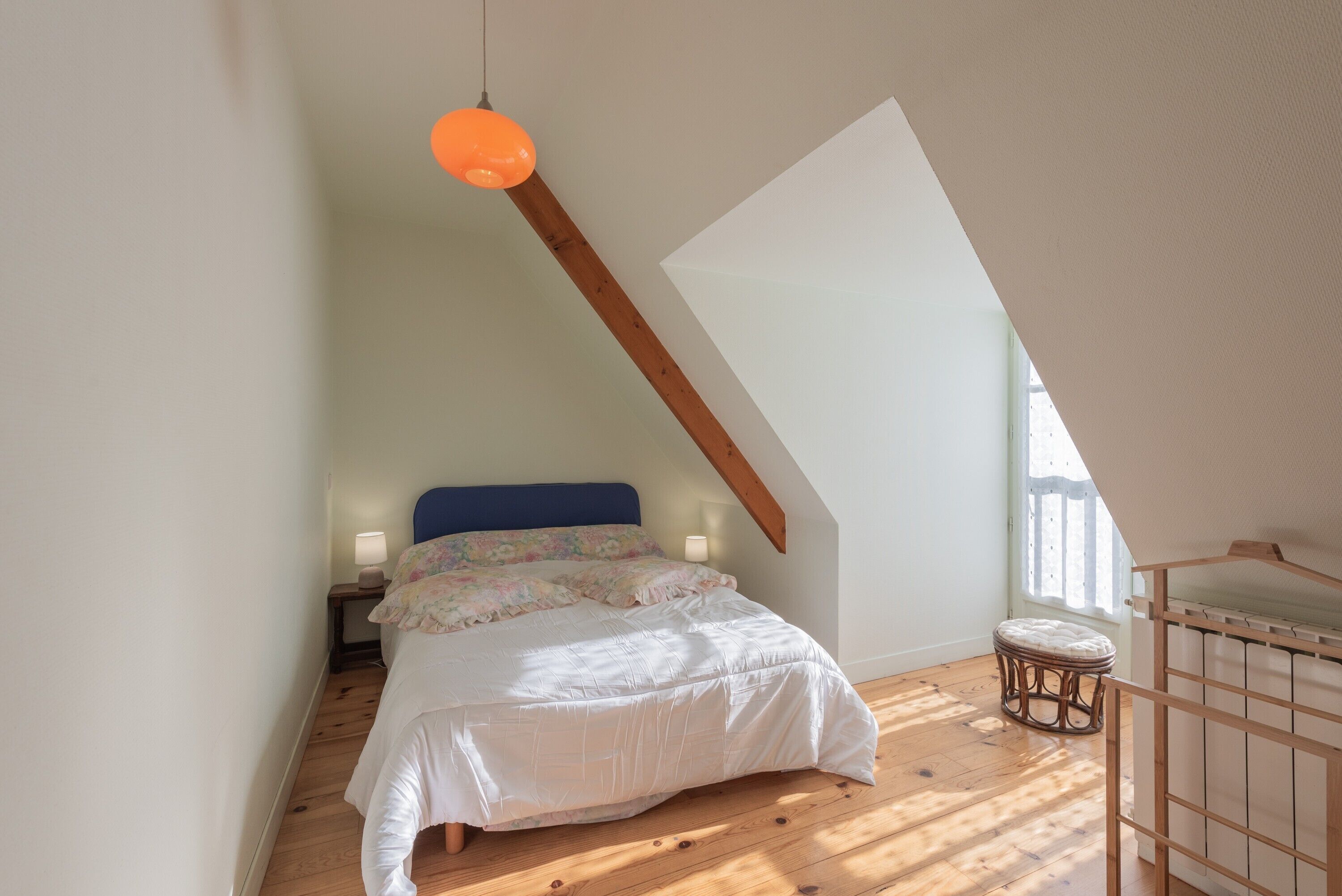 Une chambre avec un plafond en pente, un lit doté d’une tête de lit bleue, un tabouret en osier et une suspension orange.