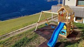 A playground with a slide, swings, and a climbing structure, set against a backdrop of mountains and a clear sky.