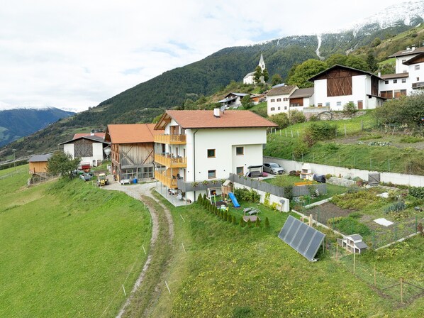 A residential area with a white house featuring a balcony, a wooden barn, and a church on a hill.