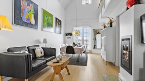 Een moderne woonkamer met een zwarte leren bank, een houten salontafel en een eethoek met een witte tafel en stoelen. Er hangen kunstwerken in lijsten aan de muren, is een open haard en is er uitzicht door een groot raam.
