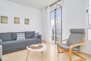 Una sala de estar moderna con un sofá gris, una silla de madera, una mesita blanca y un balcón con puertas de vidrio.