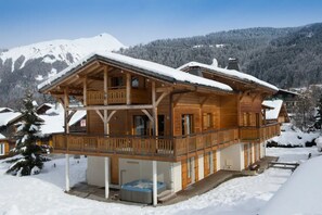 Eine Holzhütte mit Balkon und Whirlpool vor einer verschneiten Berglandschaft.