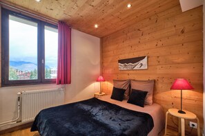 A wooden-paneled room with a bed, two lamps, a radiator, and a window with a view of mountains.
