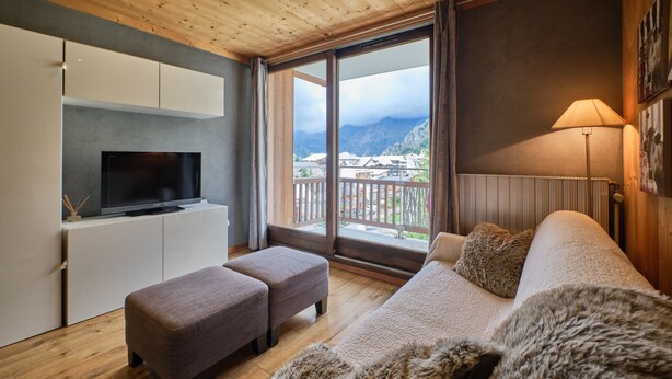 A modern room with a TV, sofa, ottoman, and a large window offering a mountain view.
