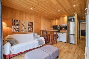 Un intérieur en bois comprenant un canapé, un pouf, un coin cuisine et des œuvres d’art murales.
