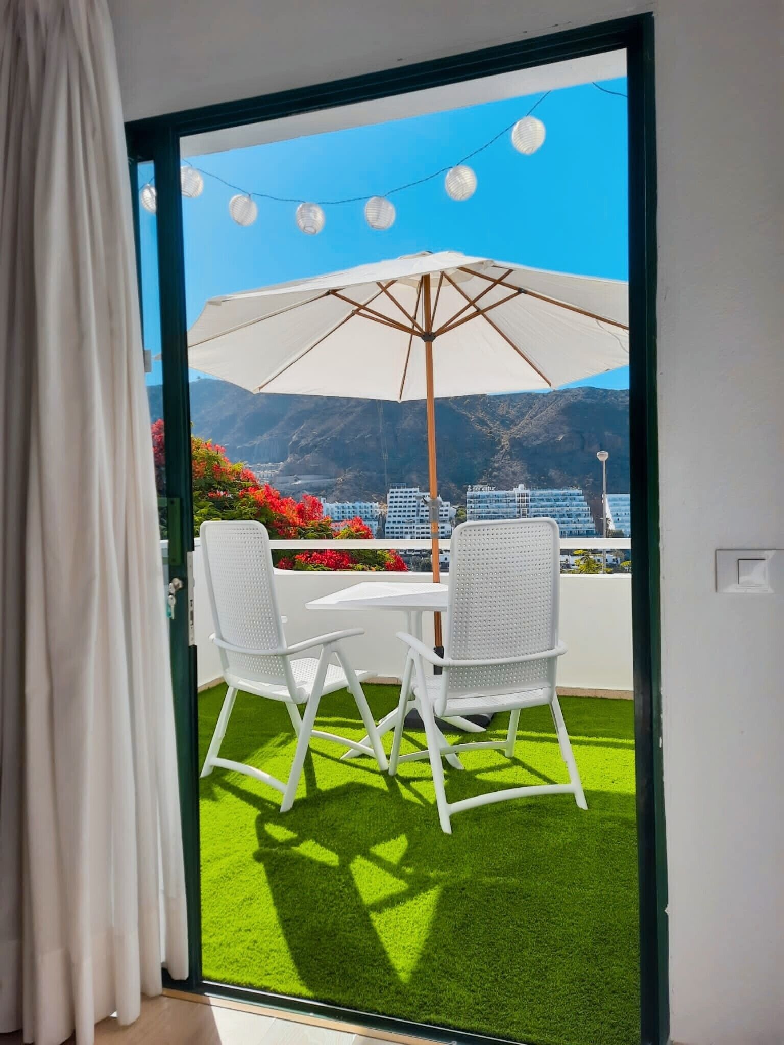 A balcony with a white table and chairs, an umbrella, and a view of mountains and buildings.