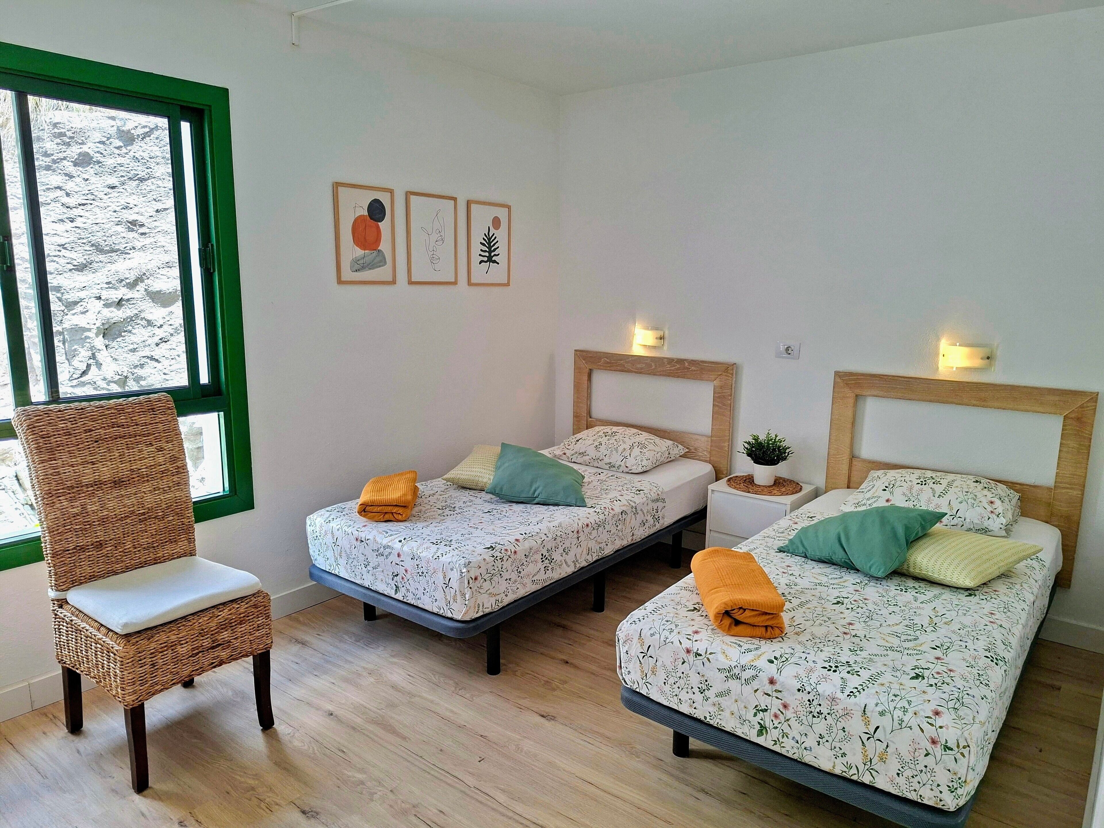 A room with two beds, a chair, a small table, and a window with a view of trees.