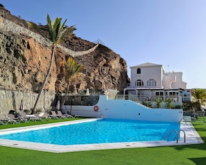 Ein Pool mit Steinmauer und Palmen, ein weißes Gebäude mit Balkon, und ein strahlend blauer Himmel.